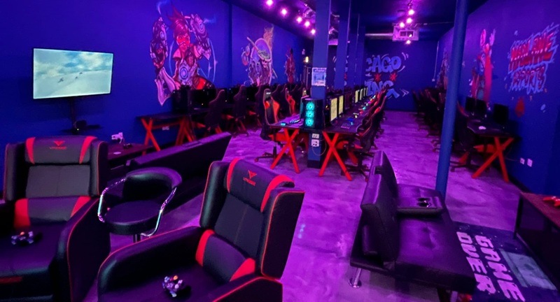 Neon-lit gaming room with red-and-black chairs, colorful wall art, LED lights, and gaming PCs in an aisle