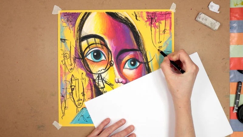 Artist hand placing blank sketch paper over colorful portrait artwork on studio worktable
