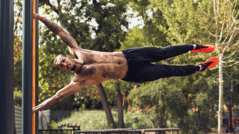 Man doing a muscle-up on a pull-up bar outdoors, shirtless with tattoo, legs extended in mid-swing