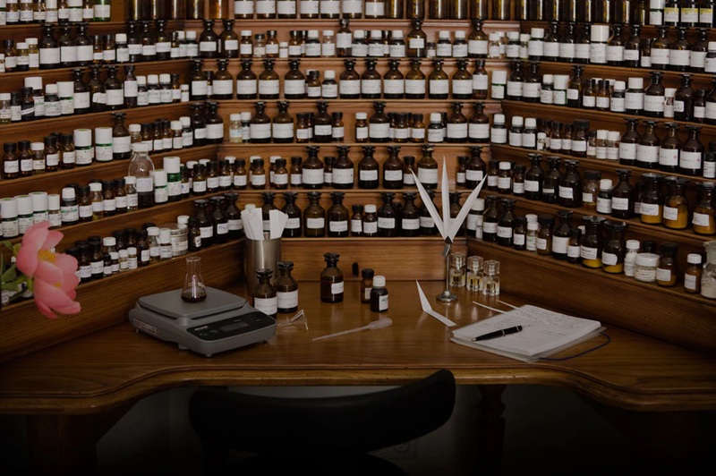 A pharmacy shelf lined with essential oil bottles, with a scale and glass vials on a wooden workbench
