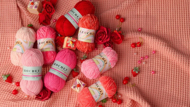 Crochet yarn skeins in red and pink arranged on a pink fabric background with scattered beads and rose decorations