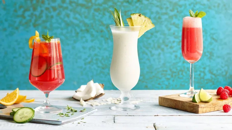 Three colorful cocktails and garnish on a wooden board: red drink, white pitcher drink, and pink berry smoothie on stem glassware