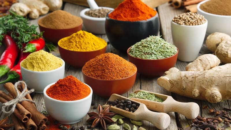 Assorted spices in bowls and wooden scoops with ginger, garlic, and herbs on a rustic table