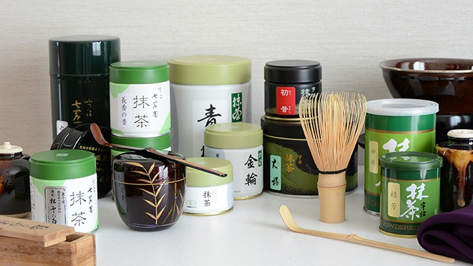 Assorted matcha and tea containers with whisk, bamboo matcha scoop, and cups on a table
