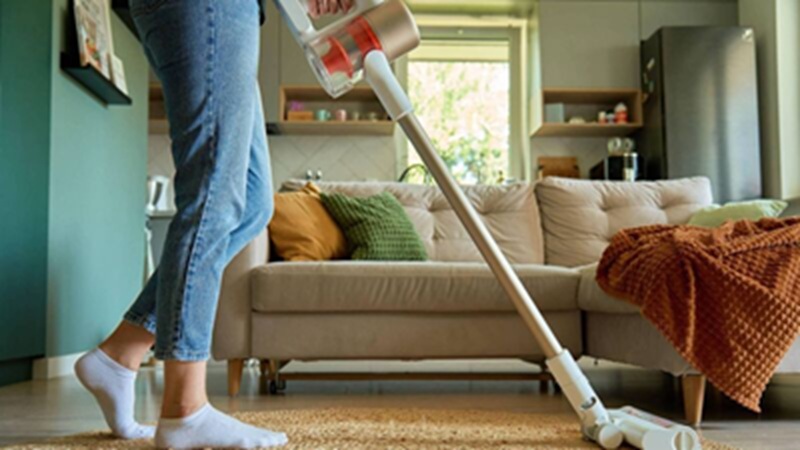 Person vacuuming a carpet in a bright living room with sofa, pillows, and window in background