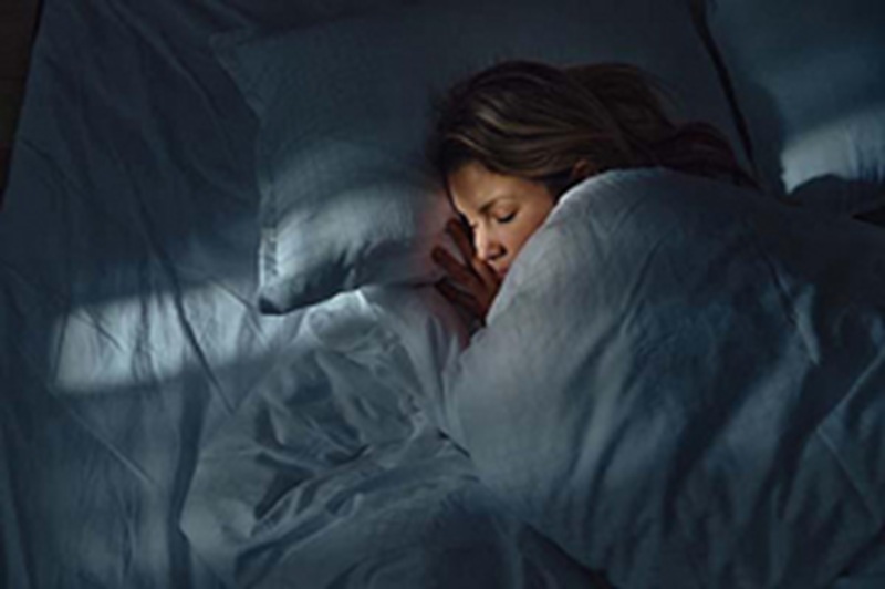 Woman sleeping under blue blanket in a dark bedroom, hugging pillow and looking weary in bed