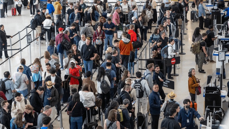 Crowd of travelers in an airport terminal with luggage and security entry gates, people waiting in lines