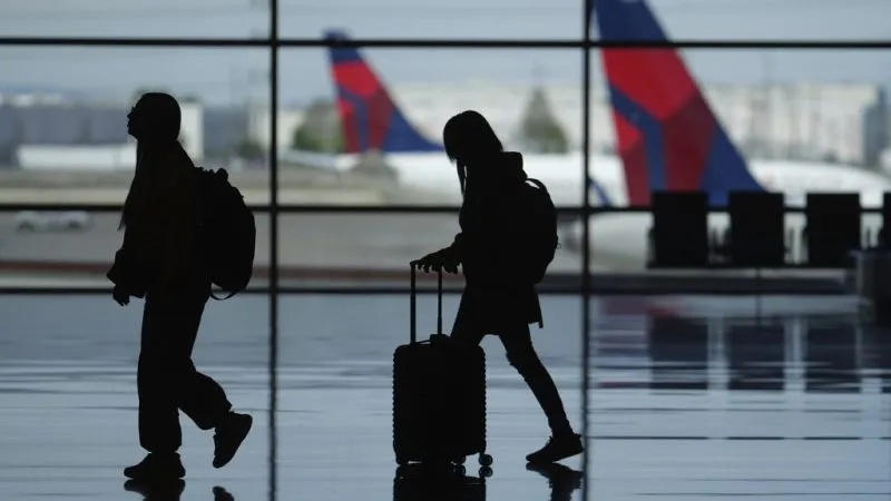 Silhouetted travelers with carry-on luggage walking past a window with distant flags and buildings