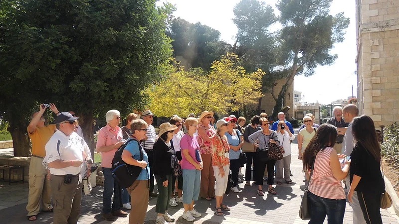 Guided tour group standing outside near trees and a stone building, listening and taking photos