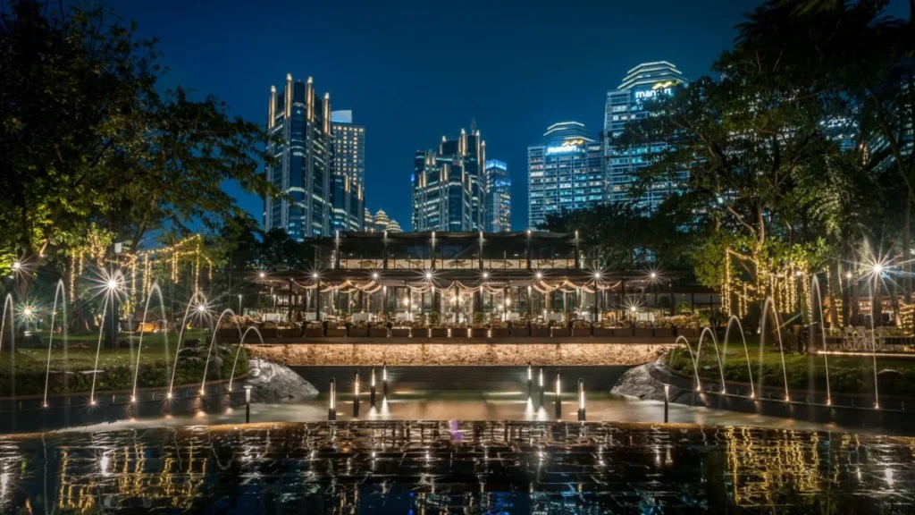 Night view of illuminated fountain with city skyline and outdoor pavilion with lights and lanterns
