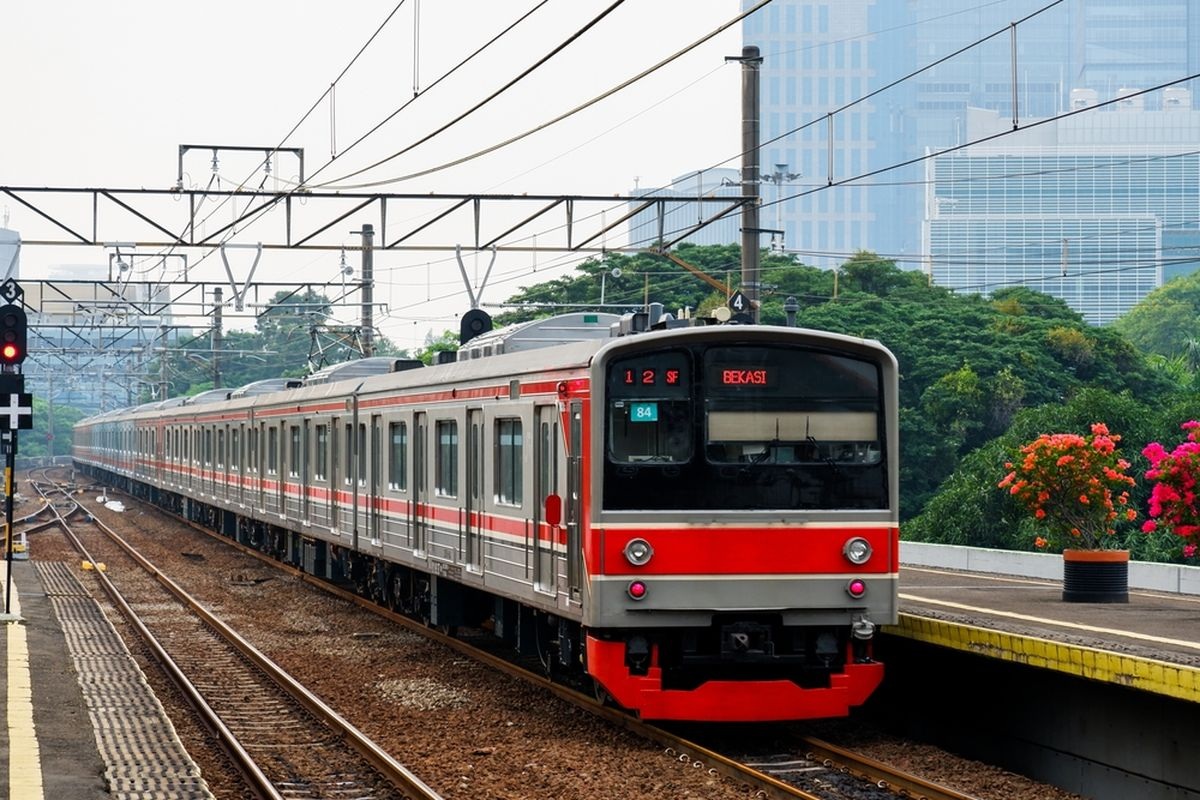 Red-and-cream commuter train arriving at station platform with skyline buildings and railway overhead wires