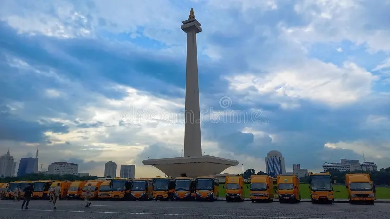 Row of city buses parked in front of a tall obelisk monument under a cloudy sky