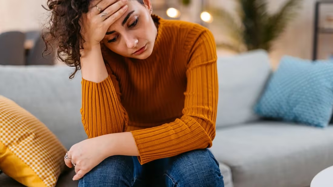 Woman in a mustard sweater sitting on a couch with her hand on her face, looking pained and holding her head