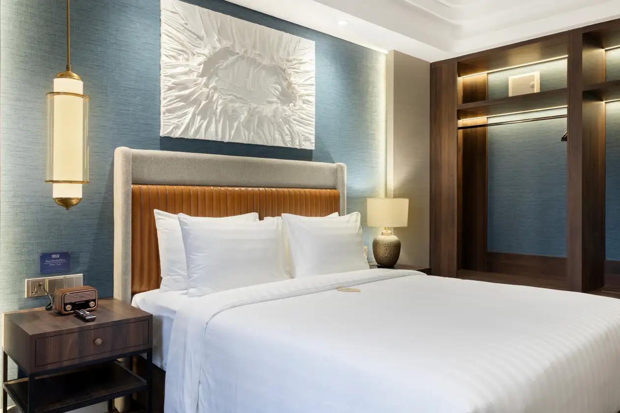 Modern hotel bedroom with tufted headboard, white bedding, bedside lamp, and framed wall art in warm lighting