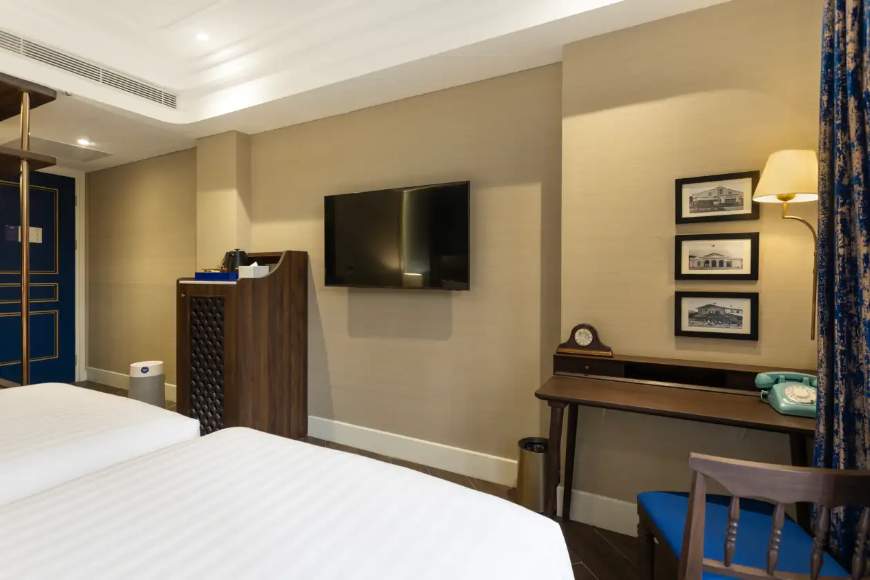 Hotel room interior with flat-screen TV, wooden reception desk, wall art frames, and bed with bedside table lamp