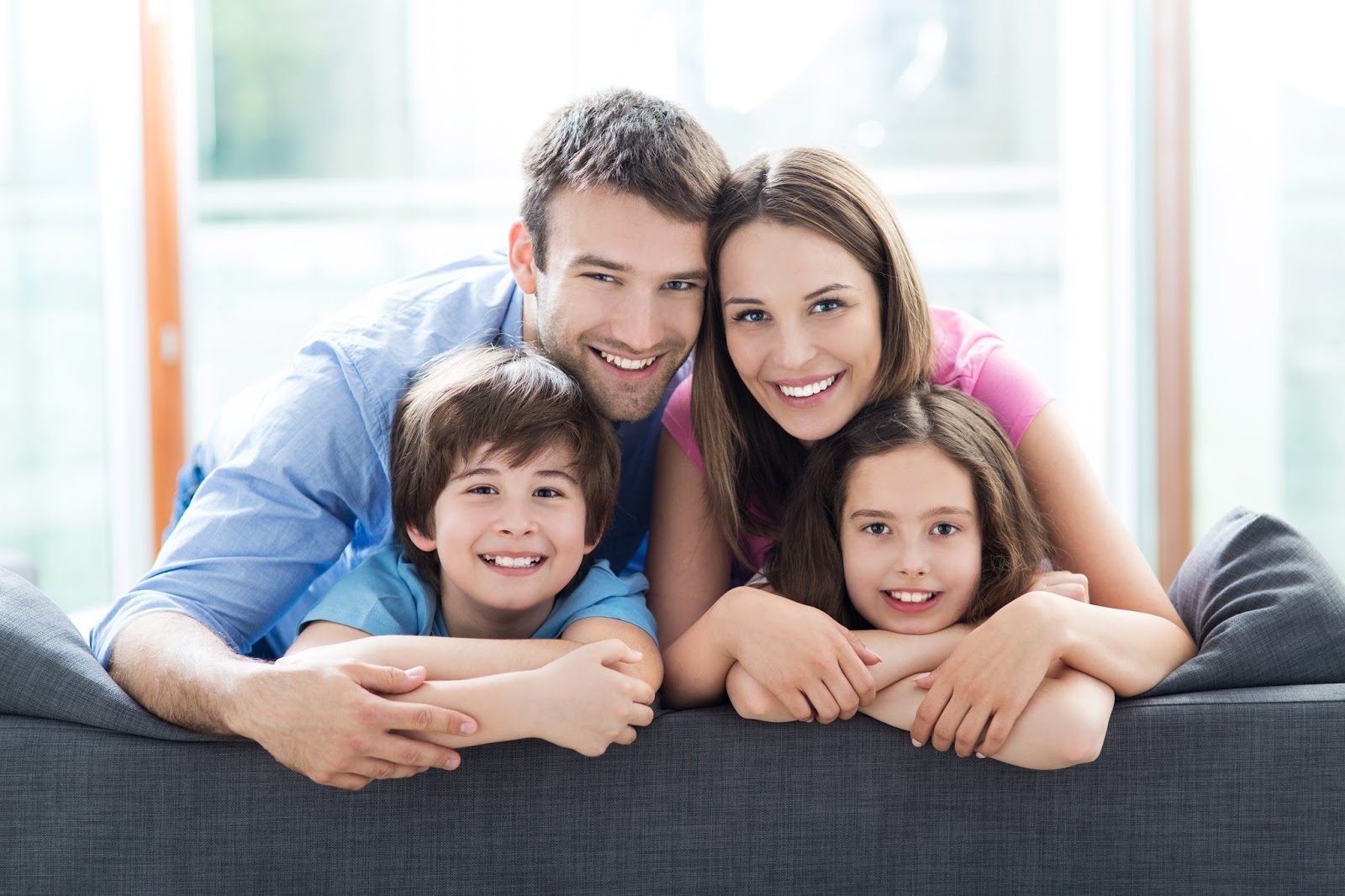 Family portrait with parents and two children smiling while leaning on a couch in bright living room
