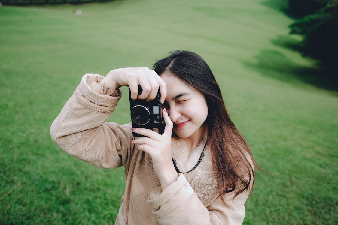 Woman taking a photo with a camera outdoors on a green lawn, smiling and holding the camera to her face