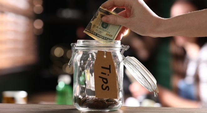 Hand dropping cash into an iDos jar labeled, with coins visible in a money jar setting