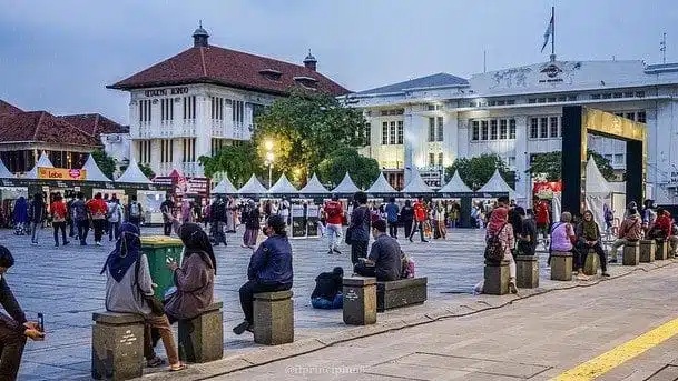 People sitting and walking at a courtyard near colonial-style buildings, umbrellas and campus tents in view
