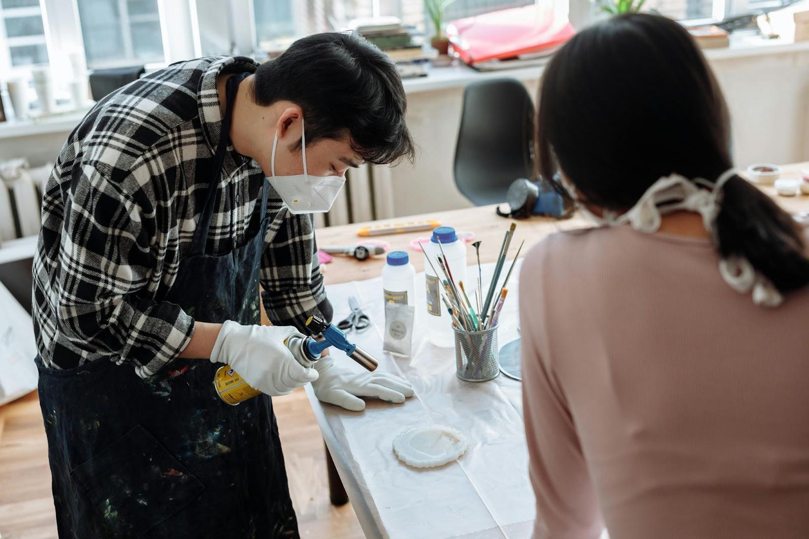 Masked students in art studio spray painting on canvas with gloves, brushes and paint bottles on table