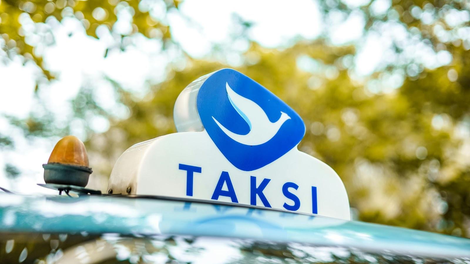 TAKSI logo on a taxi roof sign with blue flame icon, parked outdoors with trees and bokeh background