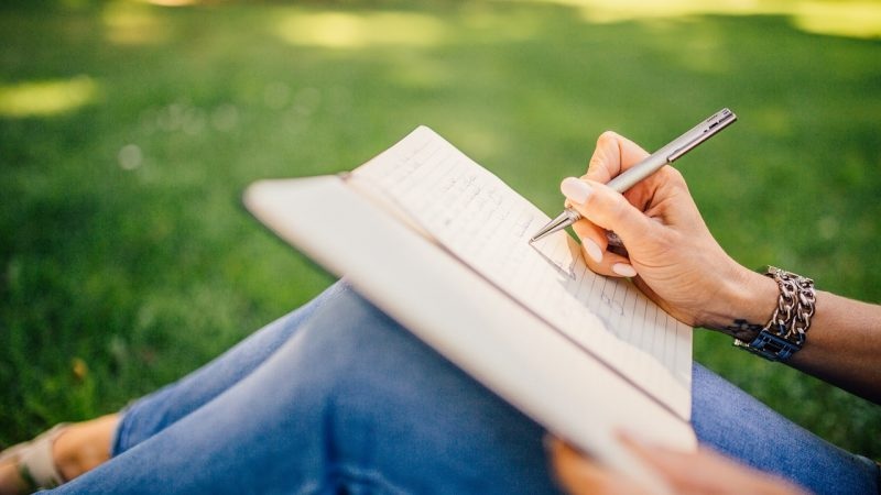 Hand writing in notebook outdoors on a grassy lawn, pen poised above open pages