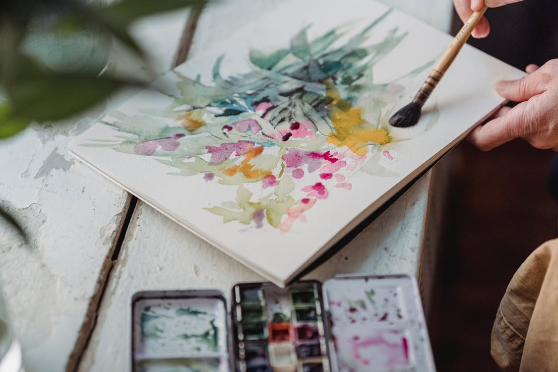Artist painting a colorful floral watercolor on paper with paintbrush, watercolors palette set nearby