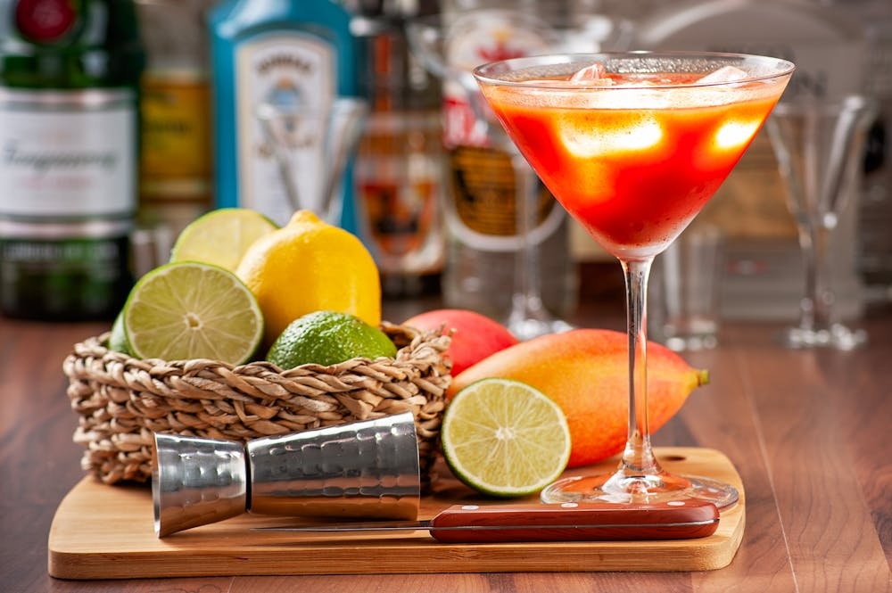 Vibrant red cocktail in martini glass on wooden tray with citrus wedges, lime, and lemon in bar background