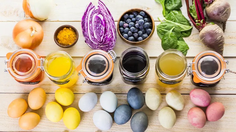 Row of fresh vegetables, herbs, eggs, and colorful jars with liquids on a light wooden kitchen table