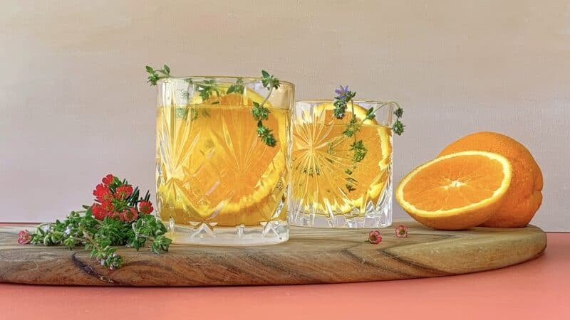 Two glasses of orange citrus drink with orange slices, fresh herbs, and flowers on a wooden table