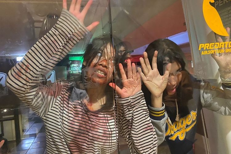 Smiling people behind glass waving hands at an airsoft shop display, with Predator sign visible in background