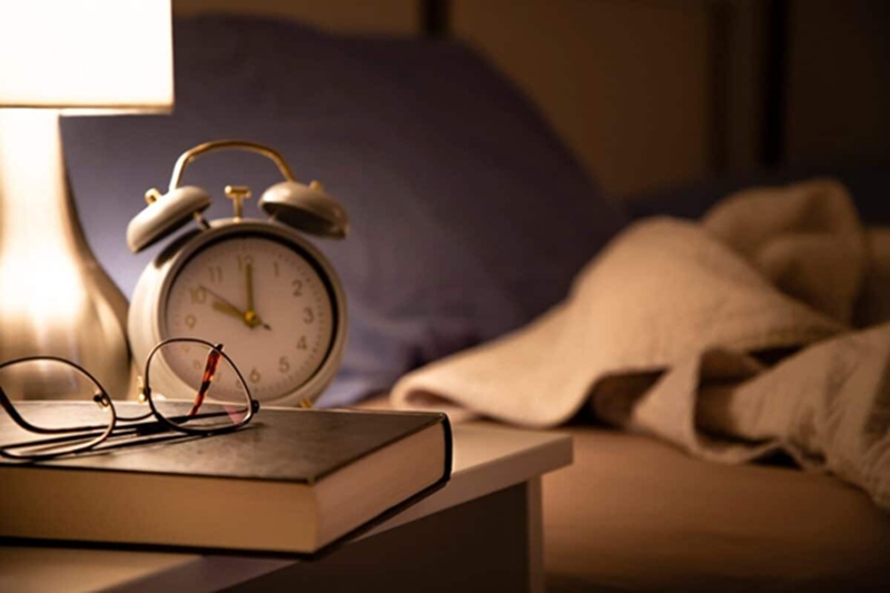 Morning alarm clock on bedside table with book and rumpled bed in softly lit room