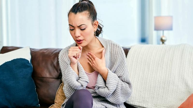 Woman sitting on a couch holding her throat, grimacing, appearing to suffer from a sore throat or pain