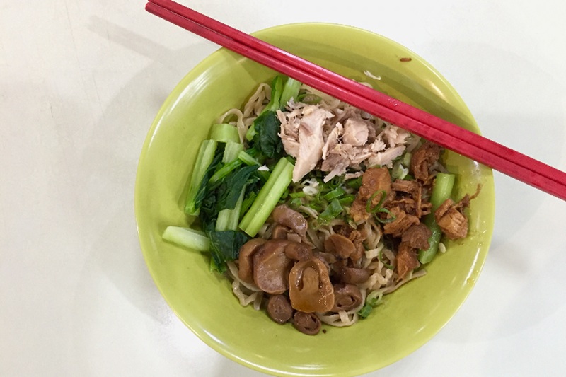 Green bowl filled with rice noodles, sliced pork, leafy greens, and vegetables with red chopsticks on top