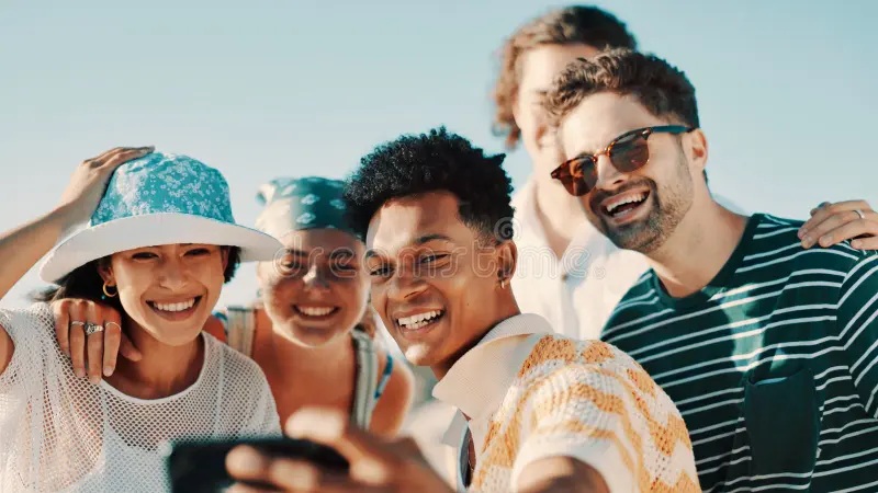 Smiling group of friends outdoors, taking a selfie with sunglasses and hats in summer light