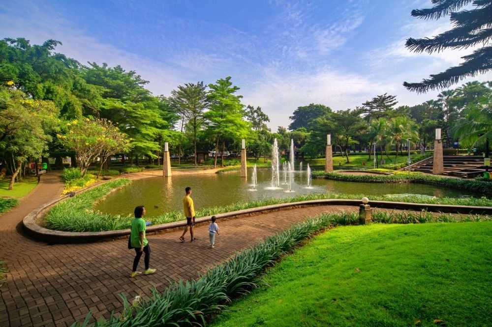 Lush garden park with fountains, brick pathways, and three people walking near a pond at dusk