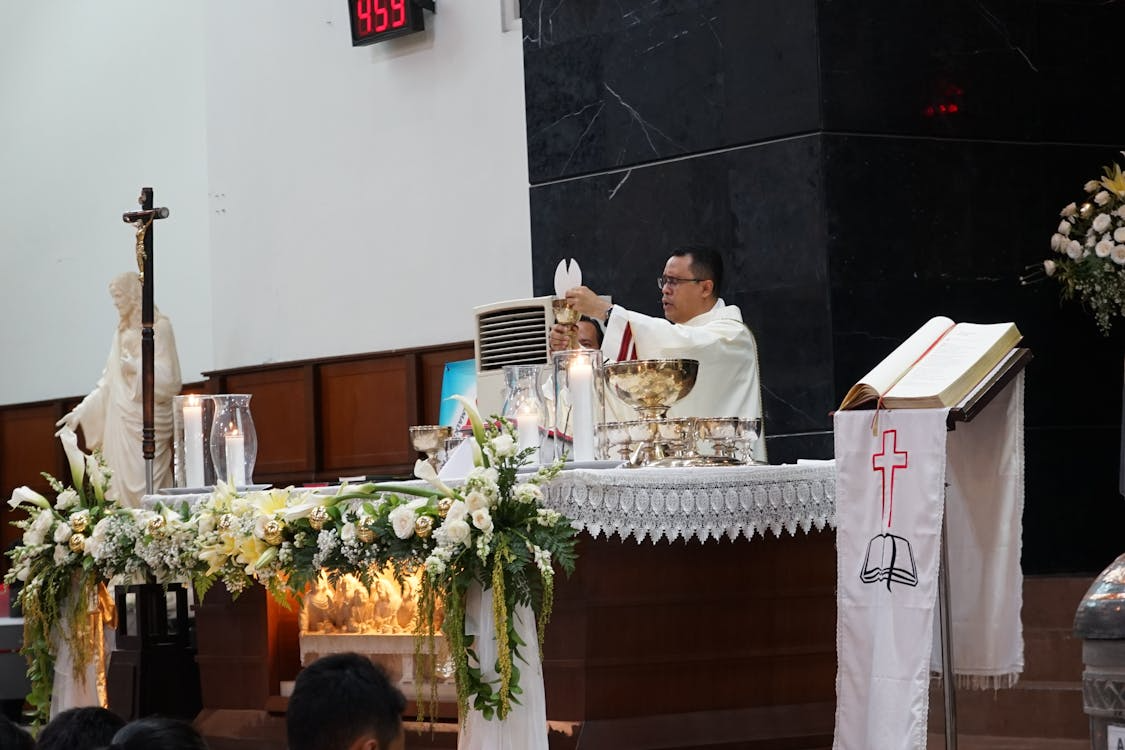 Priest in white vestments holding the communion chalice during a church service, with flowers and candles on the altar