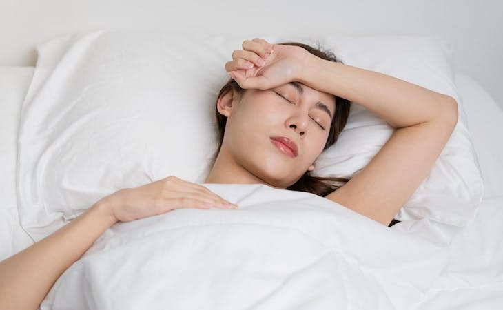 Woman sleeping in bed with eyes closed and hand on forehead, resting under white sheets