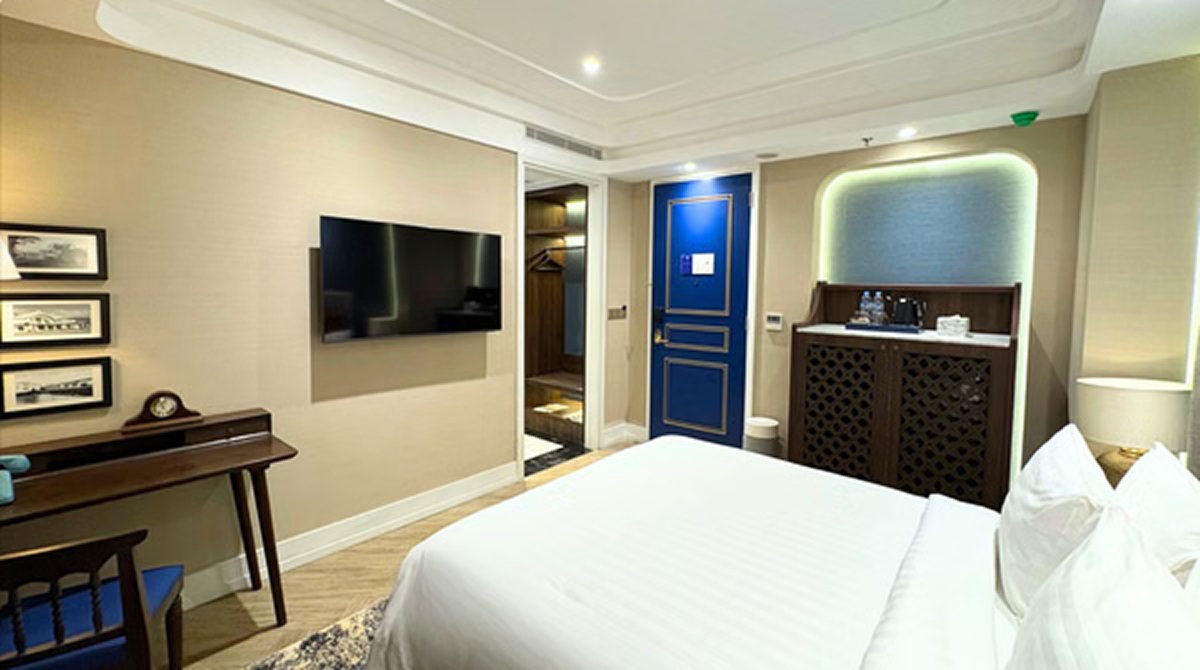 Modern hotel room with neatly made bed, wall-mounted TV, blue door, and concierge-style reception counter
