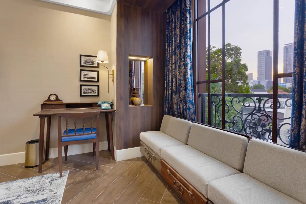 Living room corner with beige daybed, blue curtains, framed wall art, and desk near a window overlooking city skyline