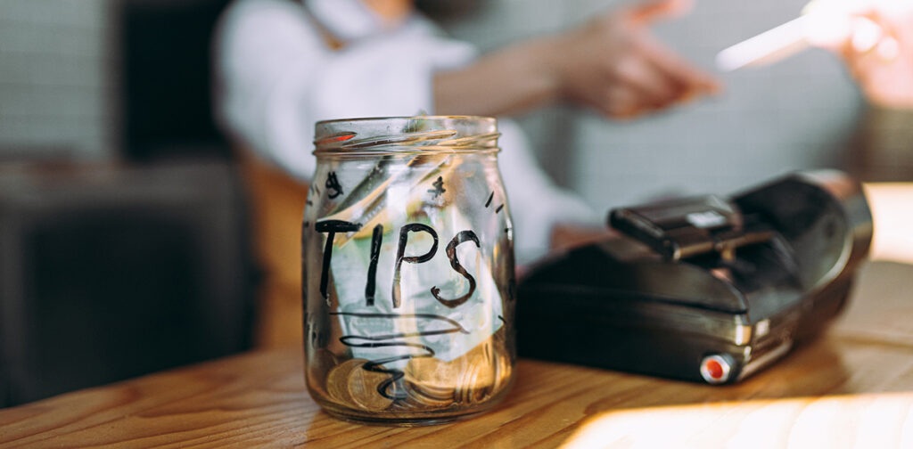 Topless glass jar labeled “TIPS” with coins and an open laptop on a wooden table, cafe setting