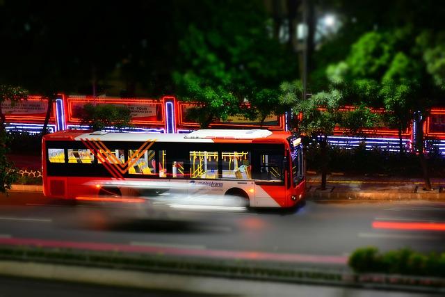 Red city bus with motion blur driving at night, illuminated street and trees in background
