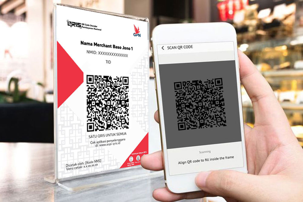 Hand holding a smartphone scanning a QR code beside a printed QR code merchant card in a store counter