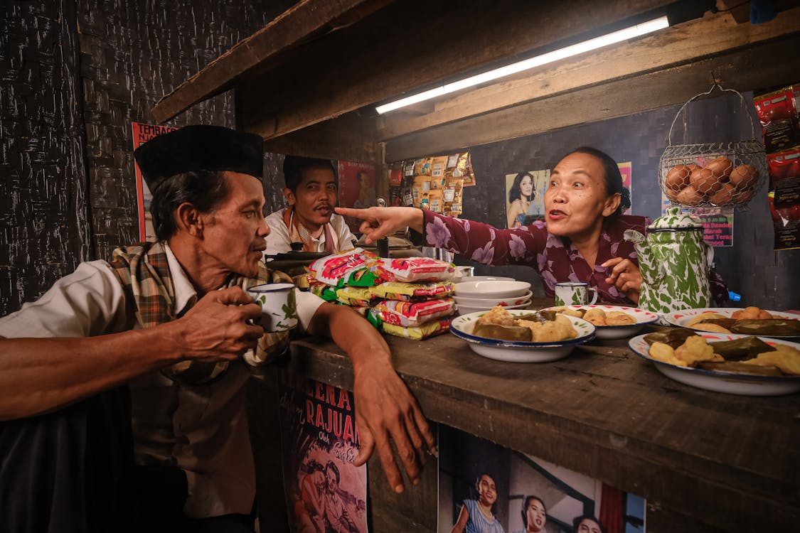 Street food stall scene with two men and an elderly woman talking while plates and packaged snacks sit on a counter