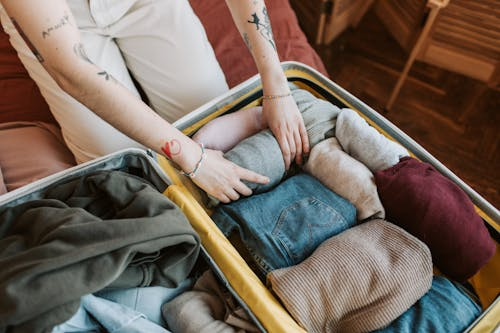 Person packing clothes and toiletries into an open suitcase, folding items on a bed with socks and jeans inside