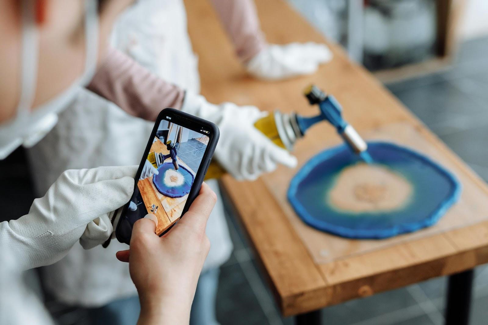 Hand holding smartphone showing a circular design for woodworking while tools and blue foam mold sit on a table