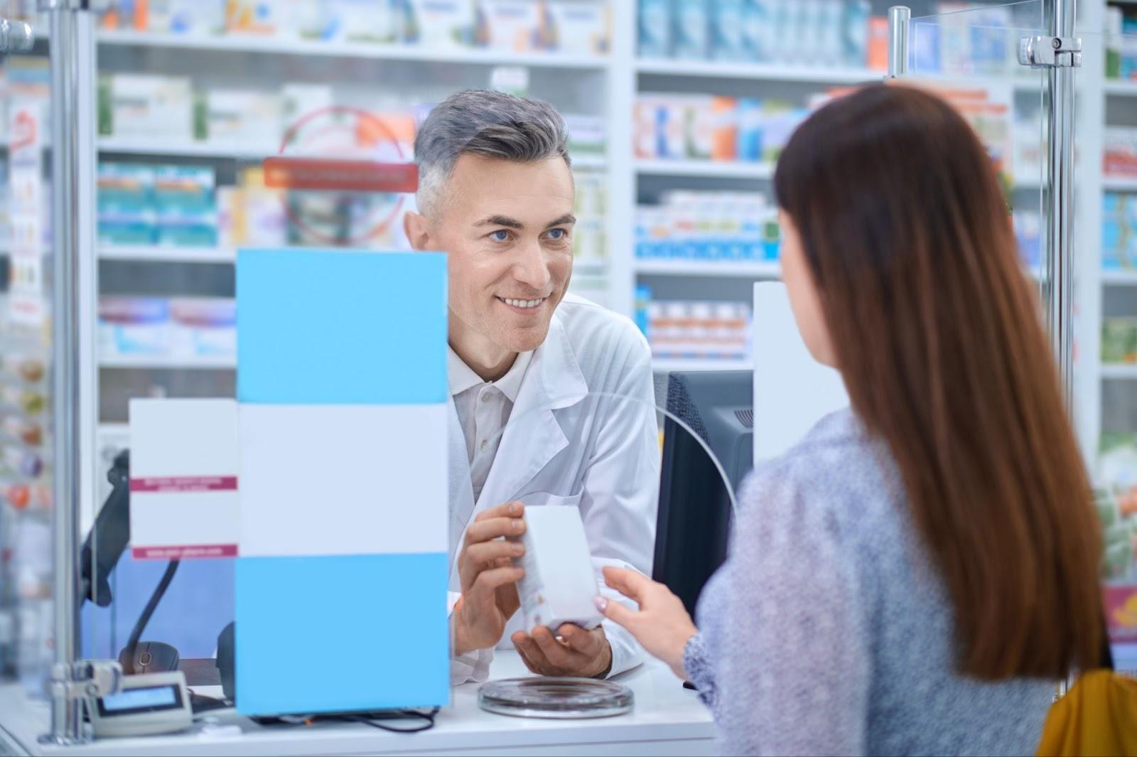 Pharmacist in pharmacy counter reviewing prescription with customer holding medication box