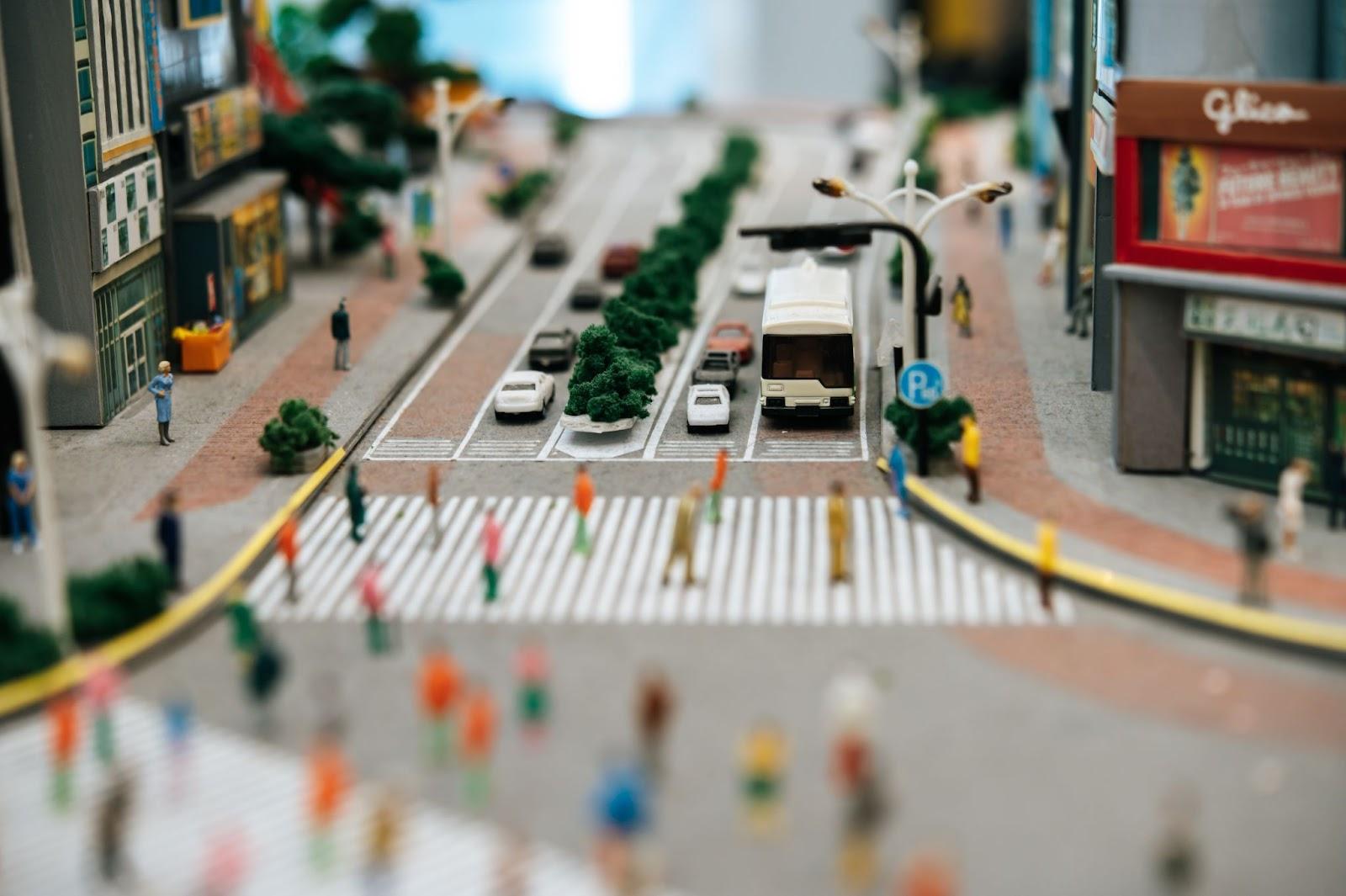 Blurred miniature city street with white delivery truck, cars, buildings, and pedestrians crossing at crosswalk