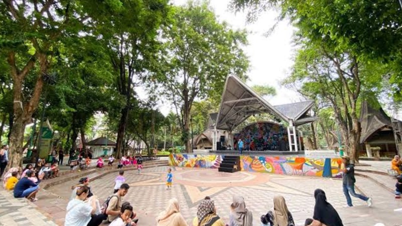 Outdoor community event in a park courtyard with trees, stage, and people walking and playing