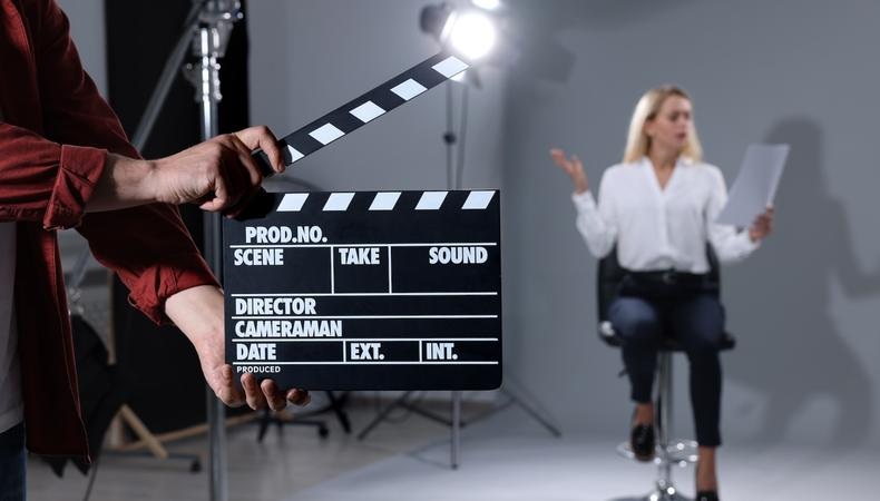 Hand holding a clapperboard labeled PROD.NO, SCENE, TAKE, SOUND with crew directing on set in background